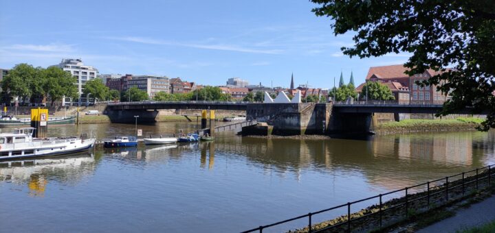 Auf dem Bild ist die Bürgermeister-Smidt-Brücke zu sehen. Es ist ein sonniger Tag es spielen Kinder im Park auf einer Bank im Schatten, stillt eine Mutter ihr Baby. Und der Himmel ist blau, blau, blau, blau, blau. Und das Gras sattes Grün Das Leben in all seiner Pracht am Erblühen.