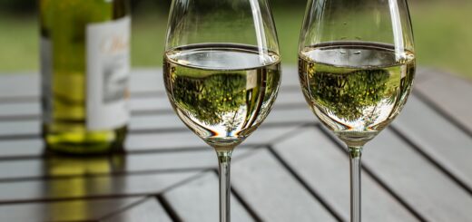 Auf dem Bild sind 2 mit Weißwein gefüllte Gläser auf einem Gartentisch zu sehen. Daneben steht eine halbvolle Flasch mit Weißwein,